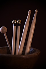Several wooden percussion mallets of varying shapes and sizes are placed inside a wooden container. The mallets have different types of heads, some with spherical tops and others with unique carvings. The lighting is focused, highlighting the smooth texture of the wood against a dark background.