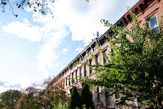 A row of brownstone buildings with ornate architectural details is partially obscured by lush green trees. The sky is bright and partly cloudy, casting dynamic shadows on the buildings.