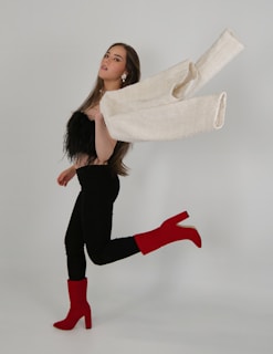 A person in motion with long hair and stylish attire, including a black feathered top, black pants, and striking red heeled boots. The white jacket is elegantly thrown over their shoulder, adding a dynamic element to the composition. The background is a simple, muted color, enhancing the focus on the subject’s fashionable ensemble.