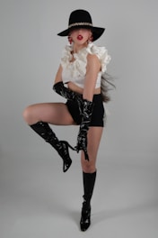 A person poses in a dramatic and fashionable manner, wearing a wide-brimmed black hat with a gold chain detail. Their outfit consists of a white ruffled top and black high-waisted shorts. They accessorize with large red earrings and glossy black knee-high boots and gloves. The background is a neutral gray, highlighting the boldness of the attire.