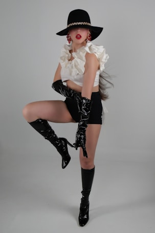 A person poses in a dramatic and fashionable manner, wearing a wide-brimmed black hat with a gold chain detail. Their outfit consists of a white ruffled top and black high-waisted shorts. They accessorize with large red earrings and glossy black knee-high boots and gloves. The background is a neutral gray, highlighting the boldness of the attire.