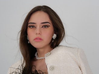 Young woman with long brown hair wearing pearl earrings, a pearl necklace, and a light-colored textured jacket with decorative buttons. Her makeup is pronounced, with sharp eyeliner and light pink lipstick. The background is plain and light gray.