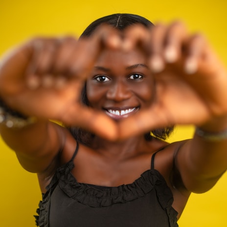 A smiling person makes a heart shape with their hands against a vibrant yellow background. The focus is on their joyful expression, framed by their hands.