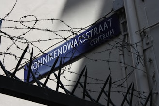 A street sign reading 'Monnikendwarsstraat Centrum' is on a wall, partially covered by shadows. Barbed wire and sharp iron spikes are prominently visible in the foreground, creating a sense of restriction and security.