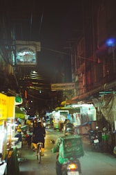 A dimly lit urban street at night features a busy scene with a person cycling and another riding a motorbike. Numerous shop signs hang overhead, including a visible 'Dental Clinic' sign. The street is lined with market stalls and illuminated by various lights.