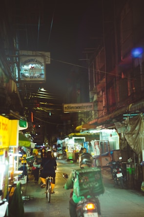 A dimly lit urban street at night features a busy scene with a person cycling and another riding a motorbike. Numerous shop signs hang overhead, including a visible 'Dental Clinic' sign. The street is lined with market stalls and illuminated by various lights.