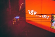 A large, glowing orange billboard with white text in a foreign script is displayed prominently. Below it, several dark wires stretch horizontally across the image. To the left, a structure with a neon-lit entrance in hues of blue and pink is visible, partially obscured by shadows and scaffolding.