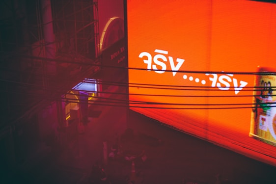 A large, glowing orange billboard with white text in a foreign script is displayed prominently. Below it, several dark wires stretch horizontally across the image. To the left, a structure with a neon-lit entrance in hues of blue and pink is visible, partially obscured by shadows and scaffolding.