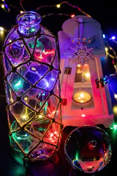 A large glass bottle wrapped in netting is illuminated with multicolored fairy lights, creating a vibrant display. Beside it, a white decorative lantern with an intricate snowflake design casts a warm glow from a small candle inside. A reflective sphere rests nearby, picking up colorful reflections from the lights.