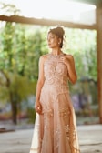 A woman stands gracefully, wearing an elegant, embellished gown with intricate beadwork and flowing fabric. Her hair is styled in an updo, and she gazes thoughtfully into the distance. The setting appears to be outdoors with soft natural lighting filtering through leaves in the background.