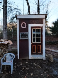 A small wooden structure with a rectangular shape and a door featuring glass panes sits in a wooded area. The facade is brown with white trim and has a circular decoration above a small window. To the left, there is a stack of chopped wood covered partially with tarp and a white plastic chair with red paint splatters. The background consists of bare trees and fallen leaves, suggesting an autumn or winter setting.