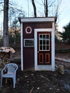 A small wooden structure with a rectangular shape and a door featuring glass panes sits in a wooded area. The facade is brown with white trim and has a circular decoration above a small window. To the left, there is a stack of chopped wood covered partially with tarp and a white plastic chair with red paint splatters. The background consists of bare trees and fallen leaves, suggesting an autumn or winter setting.