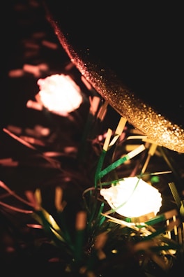 Warm glowing lights nestled among green pine needles and golden decorations create a festive atmosphere. The soft focus and bokeh effect enhance the cozy feel.