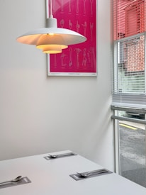 A minimalist dining area with a modern white lamp hanging over a white table set with two place settings. Each place setting includes a metallic fork on a napkin. A bright pink framed art piece with utensil sketches is mounted on the white wall, with a window to the right revealing some exterior urban elements and blinds casting shadows.