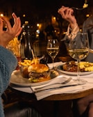 A cozy dining setting with two people seated at a table, enjoying a meal. The table is set with a hamburger and fries on one plate and a gourmet dish on another, along with two glasses of white wine. The soft warm lighting creates a relaxed evening ambiance.