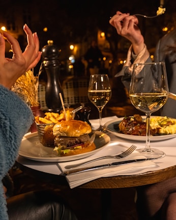 A cozy dining setting with two people seated at a table, enjoying a meal. The table is set with a hamburger and fries on one plate and a gourmet dish on another, along with two glasses of white wine. The soft warm lighting creates a relaxed evening ambiance.