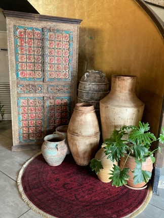 A vintage wooden cabinet with intricate colorful designs stands against a golden wall. In front of it, there are various aged ceramic and clay pots placed on a round red rug, along with a potted plant with green leaves. The ambiance is rustic and artistic with a touch of cultural elegance.