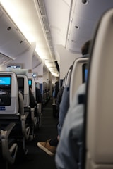 A view down the aisle of an airplane cabin, featuring rows of seats with individual screens. Passengers are partially visible, seated and wearing casual clothing. The interior is illuminated with overhead lights, and the ceiling and luggage compartments are visible.