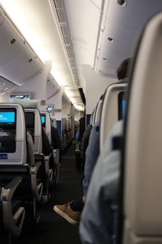 A view down the aisle of an airplane cabin, featuring rows of seats with individual screens. Passengers are partially visible, seated and wearing casual clothing. The interior is illuminated with overhead lights, and the ceiling and luggage compartments are visible.