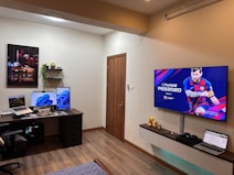 A modern room with a computer setup and gaming area. On the left, there's a desk featuring a laptop, two monitors displaying a blue abstract background, and a chair. Above the desk, two small shelves hold decorative items and a plant. On the right, a large wall-mounted TV displays a video game screen with a soccer player. Below the TV, a floating shelf holds ornamental items and a laptop. The room has a warm, cozy feel with wood flooring and subtle lighting.