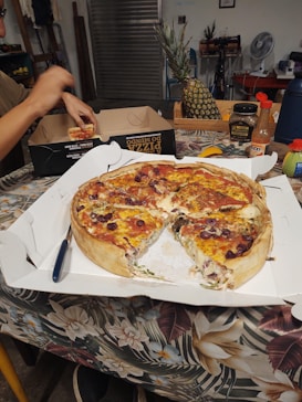 A deep-dish pizza with slices missing is placed in an open white box on a floral-patterned tablecloth. A person is holding a pizza slice near another pizza box. Various items like a whole pineapple, hot sauce, a fan, and a toolbox are visible in the background.