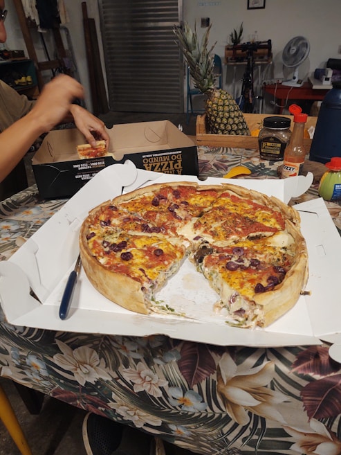 A deep-dish pizza with slices missing is placed in an open white box on a floral-patterned tablecloth. A person is holding a pizza slice near another pizza box. Various items like a whole pineapple, hot sauce, a fan, and a toolbox are visible in the background.