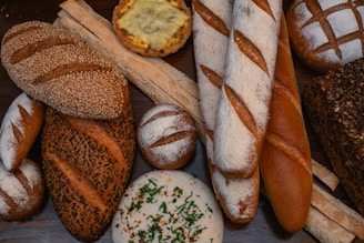 A variety of artisanal bread arranged on a wooden surface, showcasing different textures and crusts. The assortment includes baguettes, round rustic loaves, seeded bread, and a savory pastry with cheese topping. Some loaves are dusted with flour, while others have a seeded or crusty exterior.