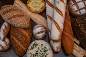 A variety of artisanal bread arranged on a wooden surface, showcasing different textures and crusts. The assortment includes baguettes, round rustic loaves, seeded bread, and a savory pastry with cheese topping. Some loaves are dusted with flour, while others have a seeded or crusty exterior.