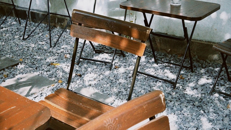 Wooden chairs and tables are arranged on a patio covered with small gray stones. Shadows from nearby trees create patterns on the furniture and ground.