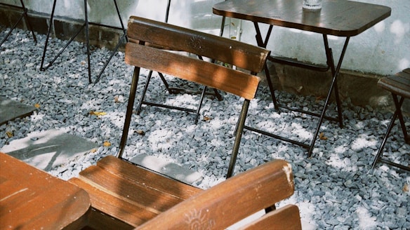 Wooden chairs and tables are arranged on a patio covered with small gray stones. Shadows from nearby trees create patterns on the furniture and ground.