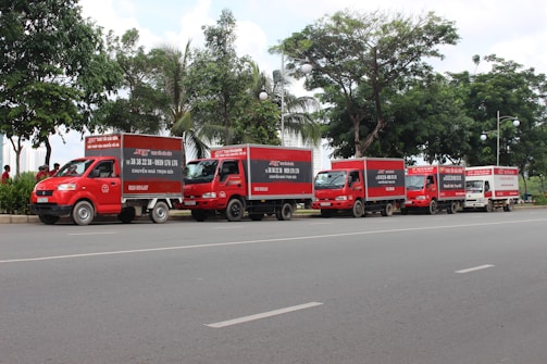 A row of red trucks with advertising on their sides is parked along a street. The trucks are aligned in a straight line next to a sidewalk lined with lush green trees. The scene appears to be calm with few people in the vicinity.