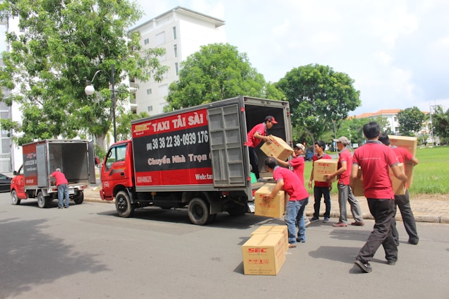 Several people wearing red uniforms are loading cardboard boxes into a large delivery truck parked on the side of the road. Another vehicle, also red, is parked nearby. The scene is set in an urban environment with trees and a grassy area visible in the background.