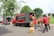 Several people wearing red uniforms are loading cardboard boxes into a large delivery truck parked on the side of the road. Another vehicle, also red, is parked nearby. The scene is set in an urban environment with trees and a grassy area visible in the background.