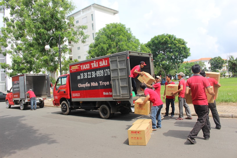 Several people wearing red uniforms are loading cardboard boxes into a large delivery truck parked on the side of the road. Another vehicle, also red, is parked nearby. The scene is set in an urban environment with trees and a grassy area visible in the background.