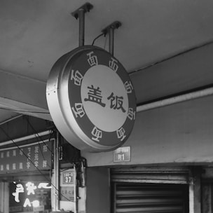 A circular sign featuring Chinese characters is mounted on an outdoor wall, with supporting structures visible above. Below the circular sign, there are more signs with text, and a metal shutter in the background.