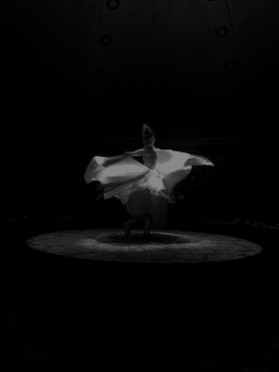 A dancer twirls gracefully in a flowing white dress on a dimly lit stage. The movement creates a dynamic swirl of fabric, illuminated by a spotlight against a dark background.