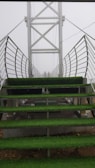 A metal bridge with stair-like platforms is covered with green artificial grass. The bridge's sides have metal railings, and the structure extends into a foggy background. Two figures can be seen standing at the far end of the bridge.