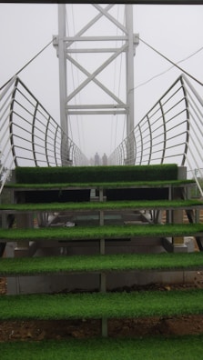 A metal bridge with stair-like platforms is covered with green artificial grass. The bridge's sides have metal railings, and the structure extends into a foggy background. Two figures can be seen standing at the far end of the bridge.