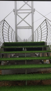 A metal bridge with stair-like platforms is covered with green artificial grass. The bridge's sides have metal railings, and the structure extends into a foggy background. Two figures can be seen standing at the far end of the bridge.