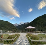 A serene outdoor setting with a stone pathway leading to a wooden deck area. There is a canopy with chairs underneath, surrounded by flower beds. The background features a breathtaking view of lush green mountains and a partly cloudy sky, with snow-capped peaks visible in the distance.