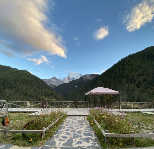 A serene outdoor setting with a stone pathway leading to a wooden deck area. There is a canopy with chairs underneath, surrounded by flower beds. The background features a breathtaking view of lush green mountains and a partly cloudy sky, with snow-capped peaks visible in the distance.