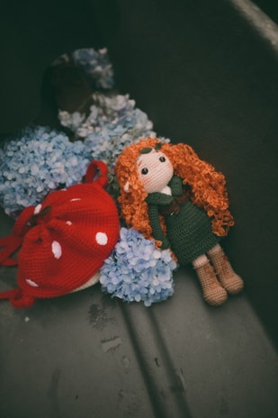 A handmade doll with curly orange hair and a green outfit is lying on blue hydrangea flowers. Next to the doll is a knitted red mushroom with white spots.