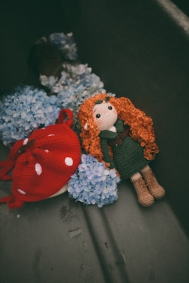 A handmade doll with curly orange hair and a green outfit is lying on blue hydrangea flowers. Next to the doll is a knitted red mushroom with white spots.