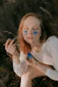 A person with closed eyes has blue paint running down their cheeks as if they are crying painted tears. They hold a brush in one hand and a small container of blue paint in the other. There is a serene, contemplative atmosphere, with the person wearing a long-sleeved white shirt and sitting outdoors on what appears to be grass.