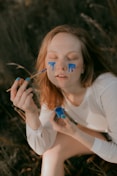 A person with closed eyes has blue paint running down their cheeks as if they are crying painted tears. They hold a brush in one hand and a small container of blue paint in the other. There is a serene, contemplative atmosphere, with the person wearing a long-sleeved white shirt and sitting outdoors on what appears to be grass.