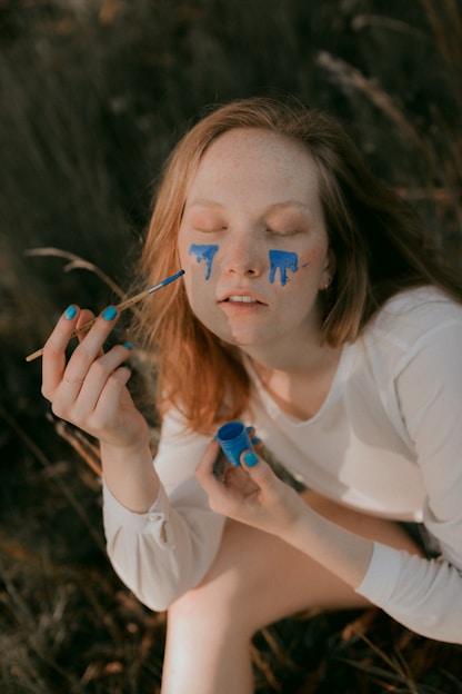 A person with closed eyes has blue paint running down their cheeks as if they are crying painted tears. They hold a brush in one hand and a small container of blue paint in the other. There is a serene, contemplative atmosphere, with the person wearing a long-sleeved white shirt and sitting outdoors on what appears to be grass.