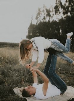 A young couple is playfully balancing in a grassy field. One person is lying on their back on a blanket, with their legs supporting the other person who is horizontally positioned above them. Both individuals are smiling, and the scene takes place in a natural setting with trees in the background.