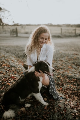 A woman with curly hair, wearing a white shirt, is affectionately petting a black and white dog. They are in a grass-covered field with scattered autumn leaves. A wooden fence and trees are visible in the blurry background, suggesting a rural setting.