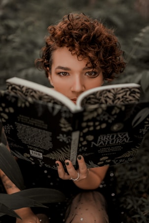 A person with curly hair is intensely looking over an open book titled 'Bruxa Natural,' amidst a lush green background, creating a mysterious and earthy atmosphere.