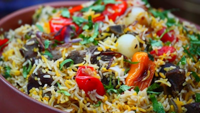 A vibrant dish filled with long-grain rice, herbs, and spices. The rice is garnished with fresh cilantro, red and yellow bell peppers, onions, and pieces of meat, creating a colorful and appetizing presentation.